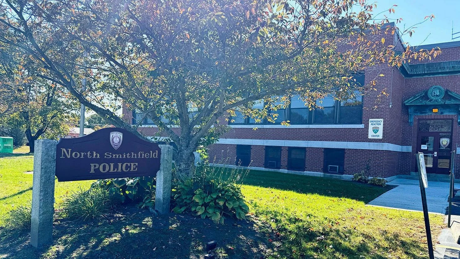 A sign for North Smithfield Police in front of a brick police station building with a tree and greenery nearby, and a sidewalk leading to the entrance.