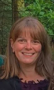 A woman with shoulder-length light brown hair and bangs, smiling outdoors in front of green trees.