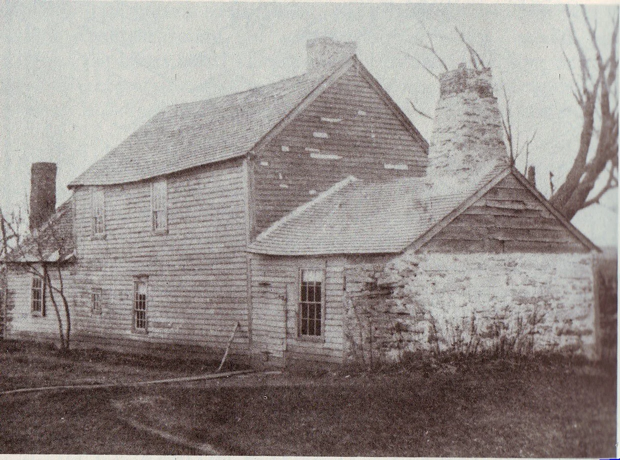 An old, weathered wooden house with a steep roof and a tall brick chimney, surrounded by bare trees, in a black-and-white photograph.