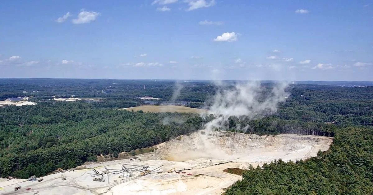 An open quarry with steaming hot surface, surrounded by green forests under a partly cloudy sky.