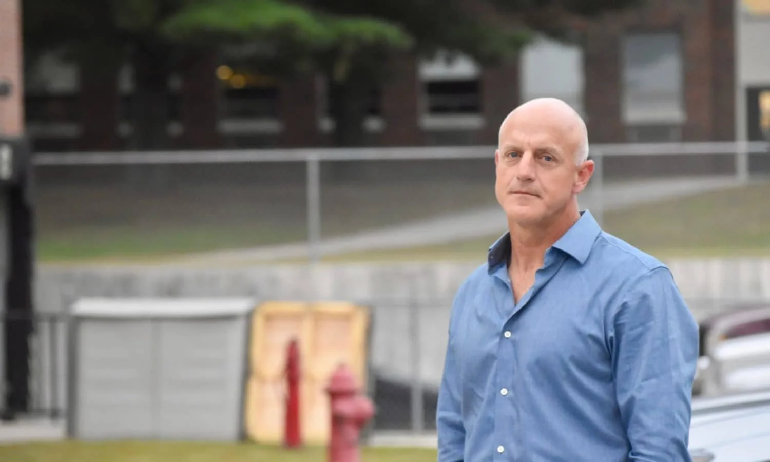 A bald man in a blue button-up shirt standing outdoors in front of a blurred urban background.
