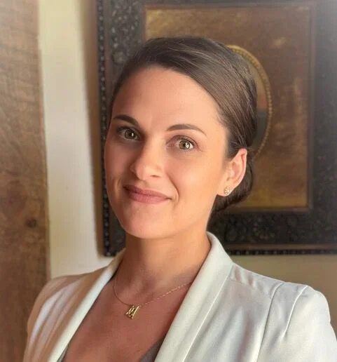 A woman with brown hair tied back, wearing a white blazer and a gold necklace, standing indoors with a framed artwork and a wooden wall in the background.