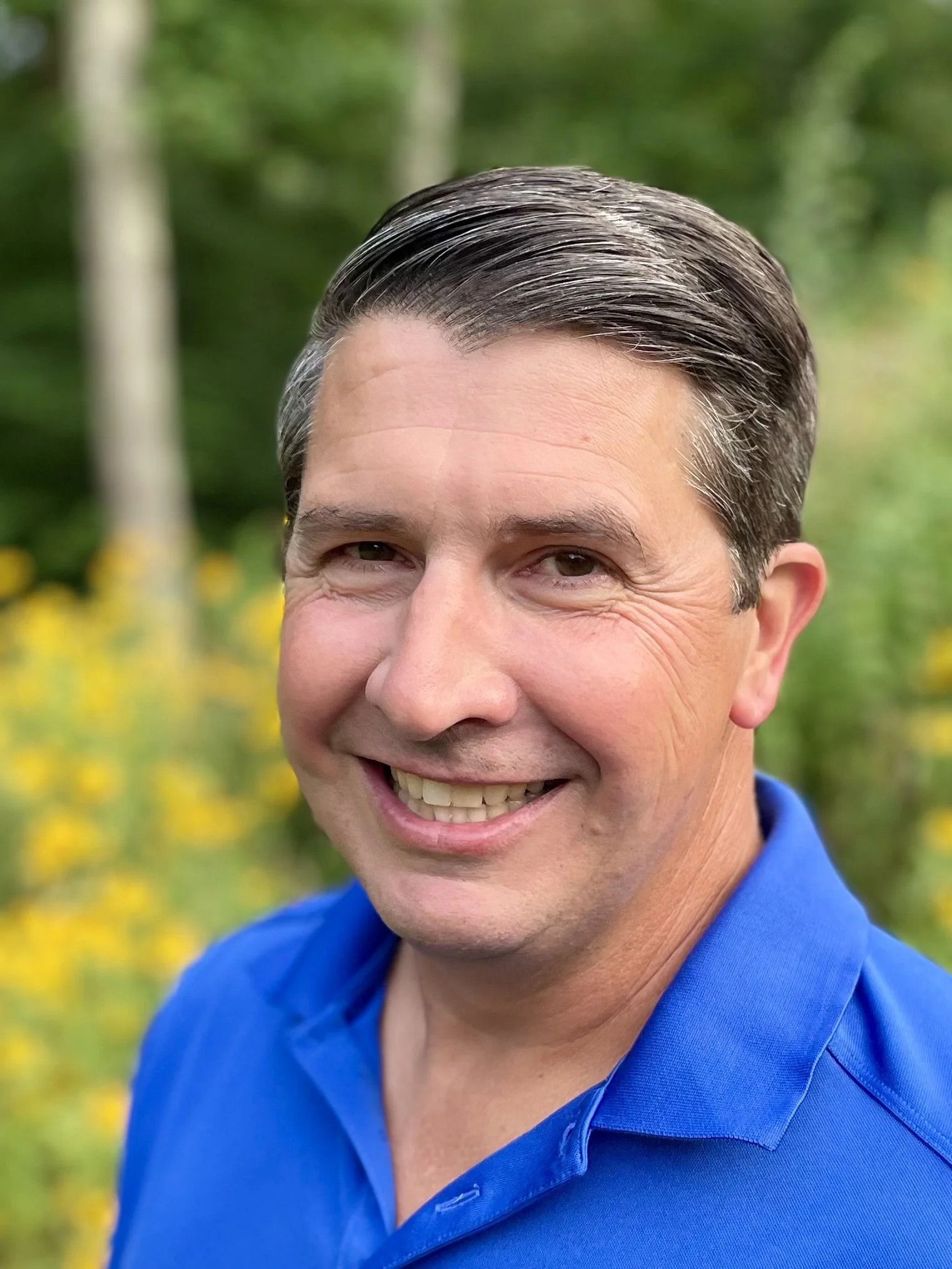 A smiling man with short dark hair, wearing a blue shirt, outdoors with green trees and yellow flowers in the background.