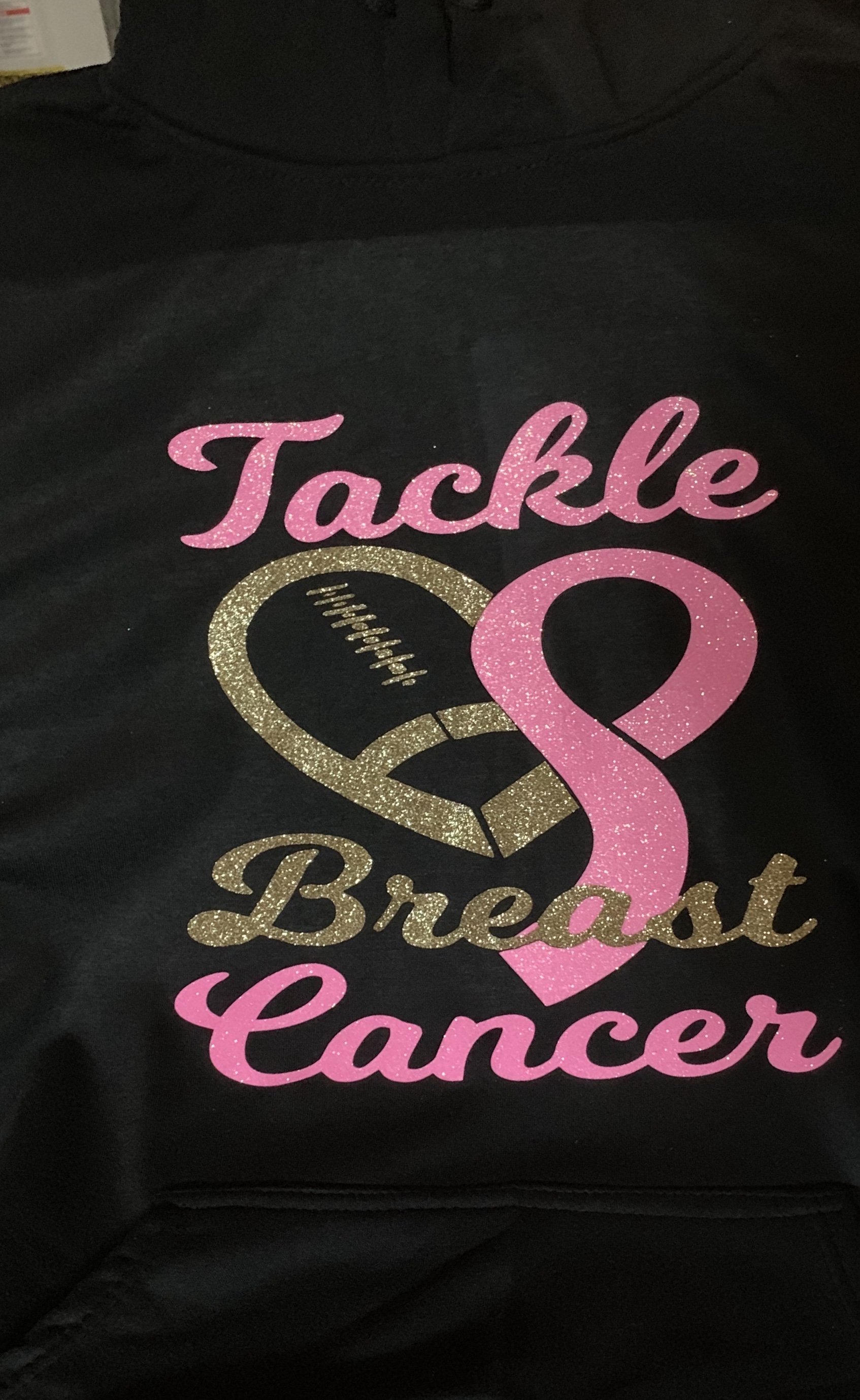 Black shirt with pink, gold, and white glitter text and graphics supporting breast cancer awareness. The design includes a pink ribbon, a football with stitching, and the words "Tackle Breast Cancer."