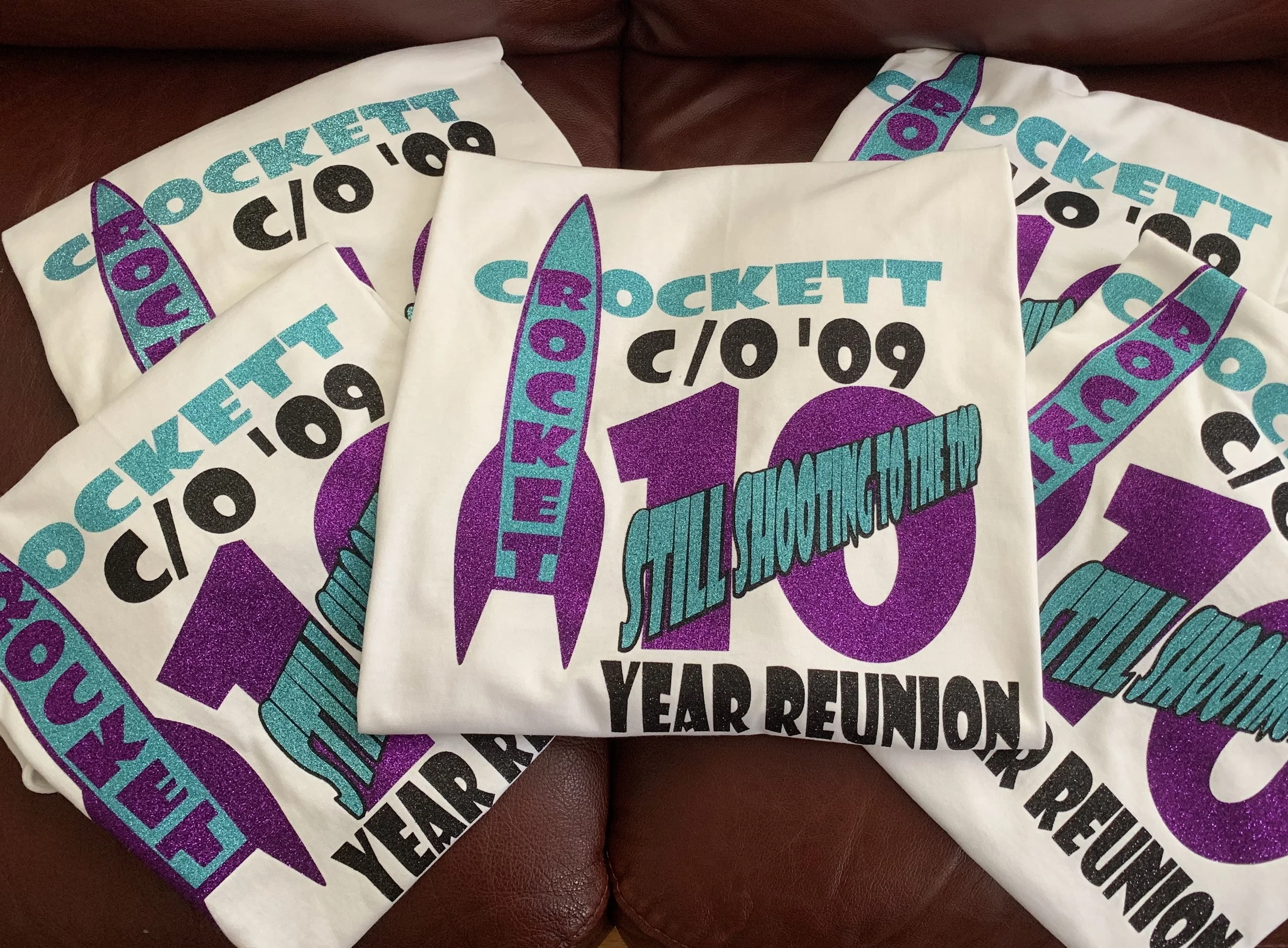 Five white T-shirts with colorful event graphics arranged on a brown leather surface. The graphic includes a large purple rocket with a teal flame, the text 'Crockett c/o '09', 'Still Shooting to the End', and mentions a 10-year reunion.