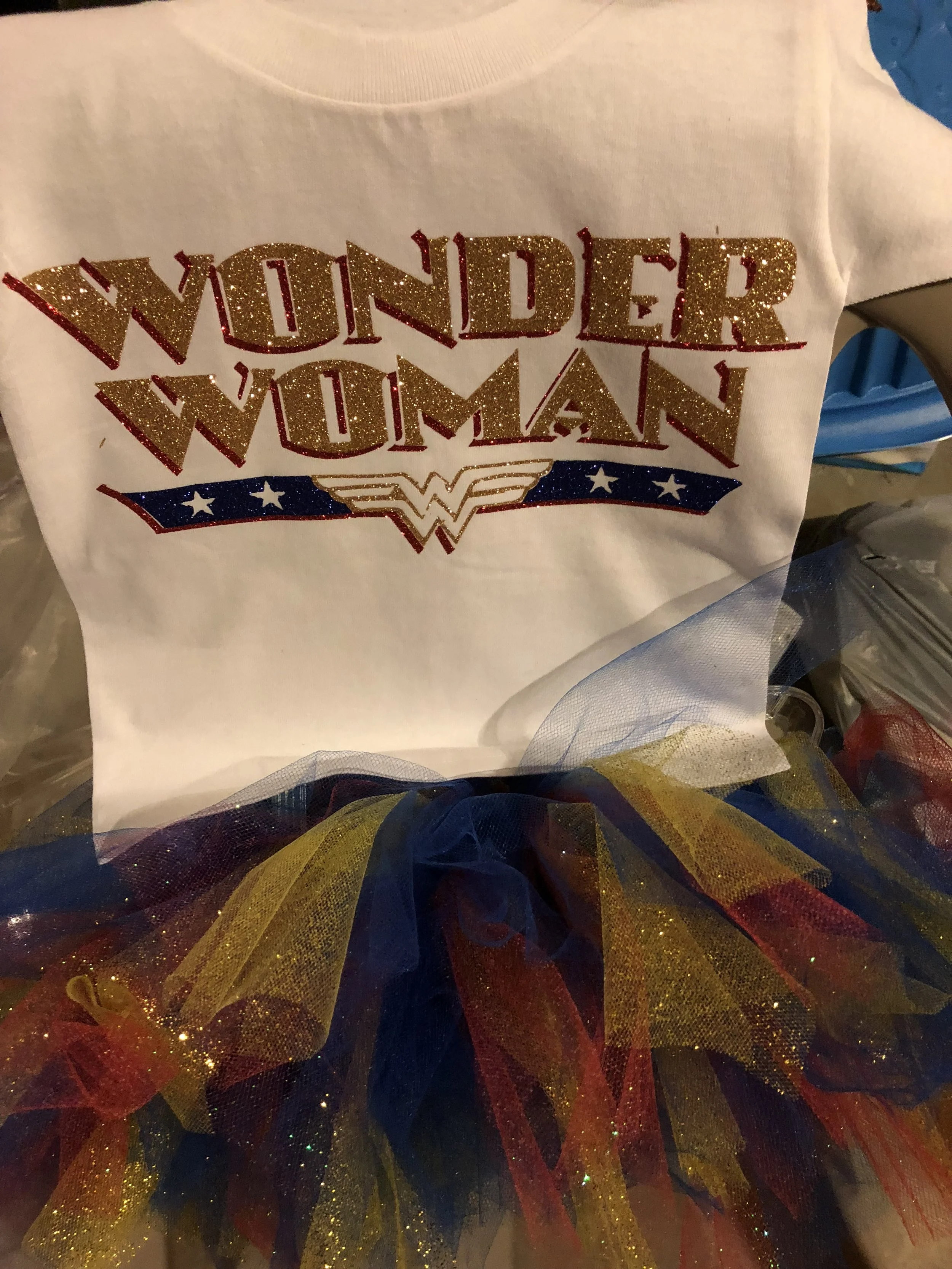 A Wonder Woman-themed t-shirt with glittery gold and red text reading 'Wonder Woman' and decorative elements, with multicolored tulle fabric underneath.