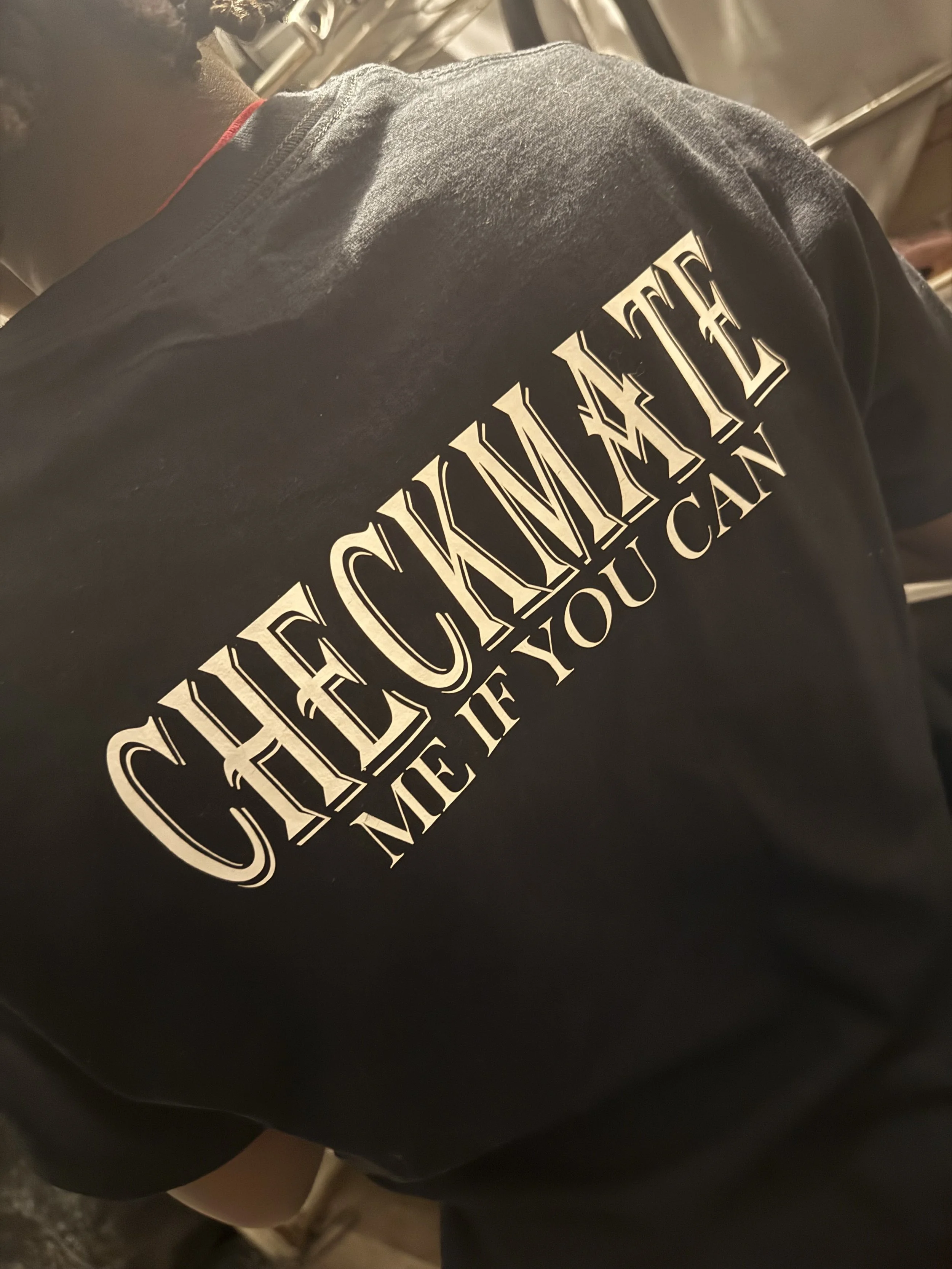 Back of a person's black t-shirt with the phrase "Checkmate" in large, decorative letters and the words "Me If You Can" below.