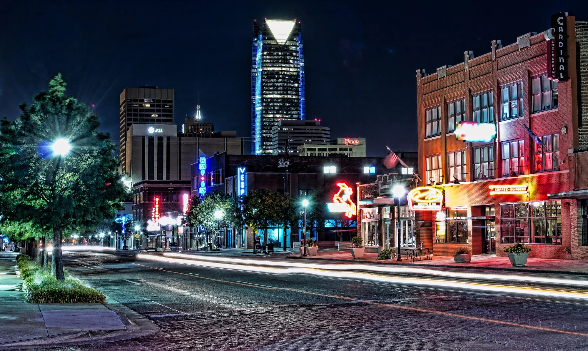 Nighttime city street scene with illuminated neon signs on buildings, a tall skyscraper in the background, and light trails from passing vehicles.