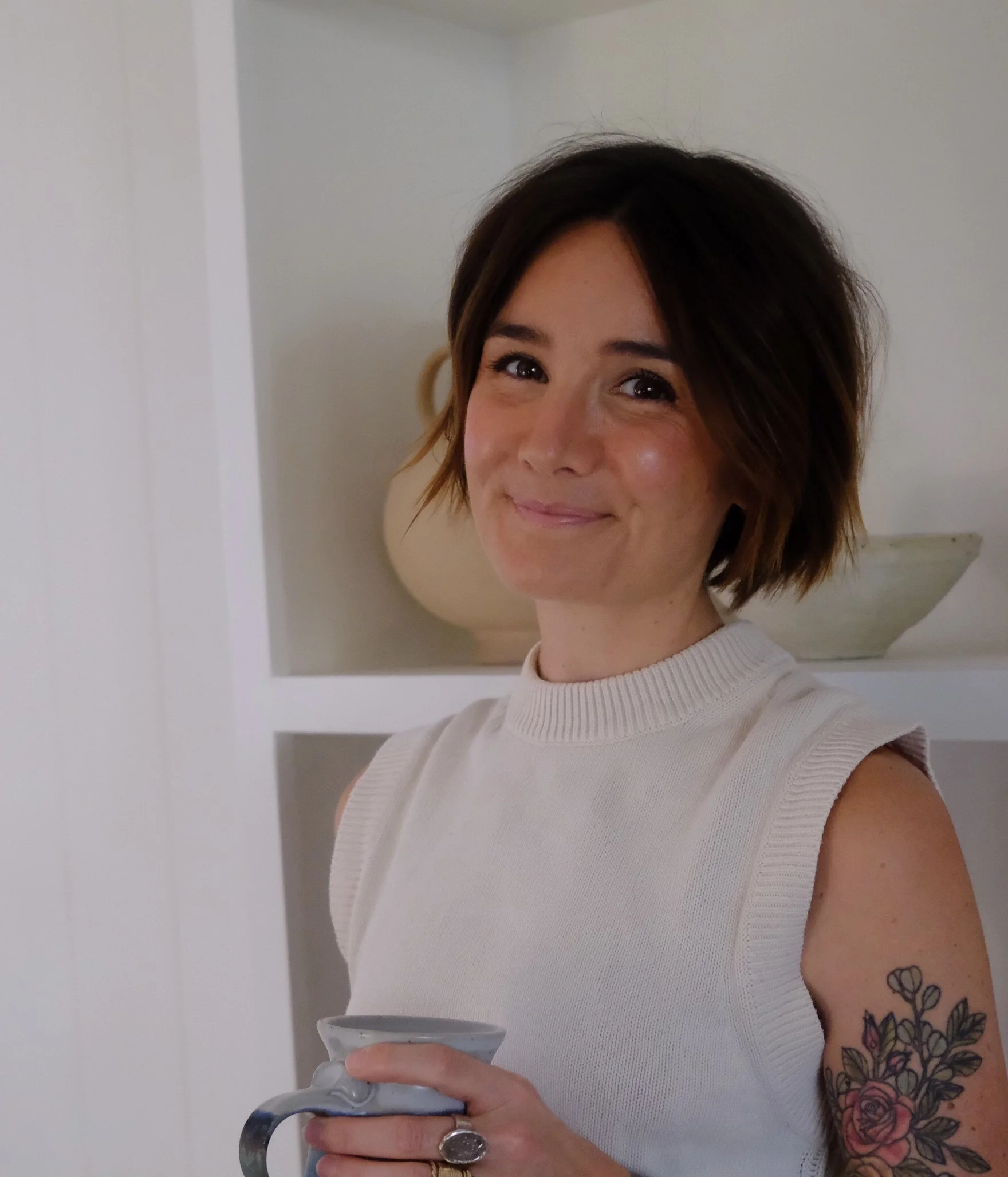 A woman with short brown hair, wearing a white ribbed sleeveless top, holds a mug and smiles at the camera. She has tattoos on her left arm, including flowers and leaves.