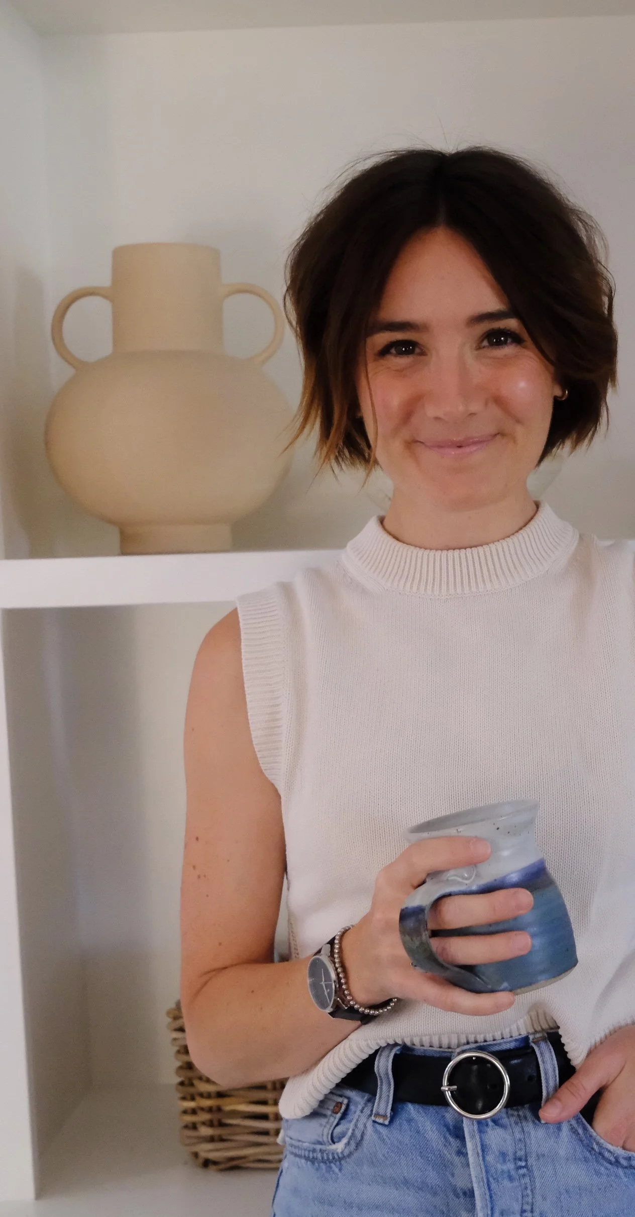 A woman with short dark hair smiling, wearing a sleeveless white top, holding a blue and gray ceramic mug, standing in front of a white shelf with a decorative beige vase.
