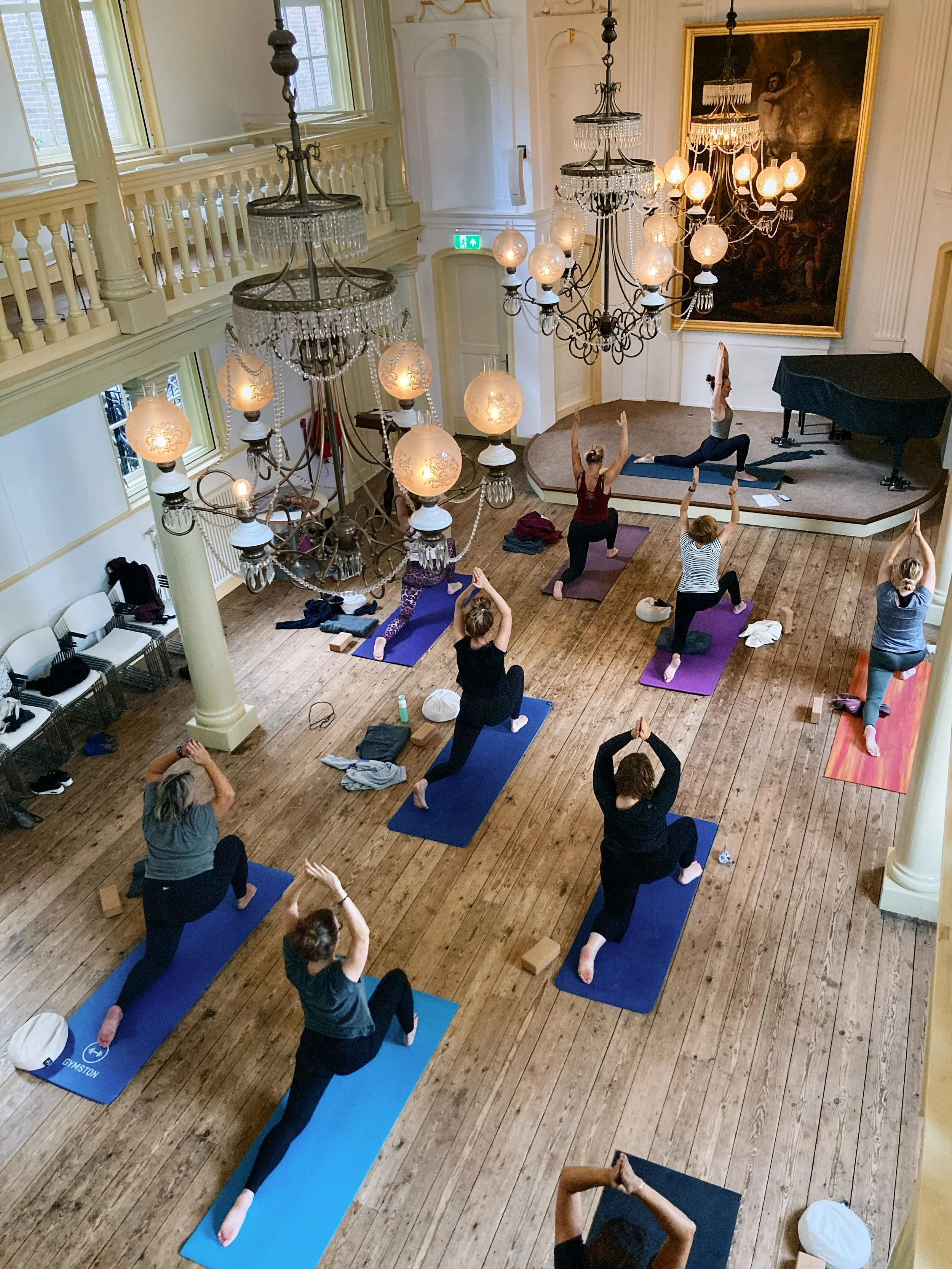 People participating in a yoga class in a grand, elegant room with chandeliers, large painting, and wooden floors.