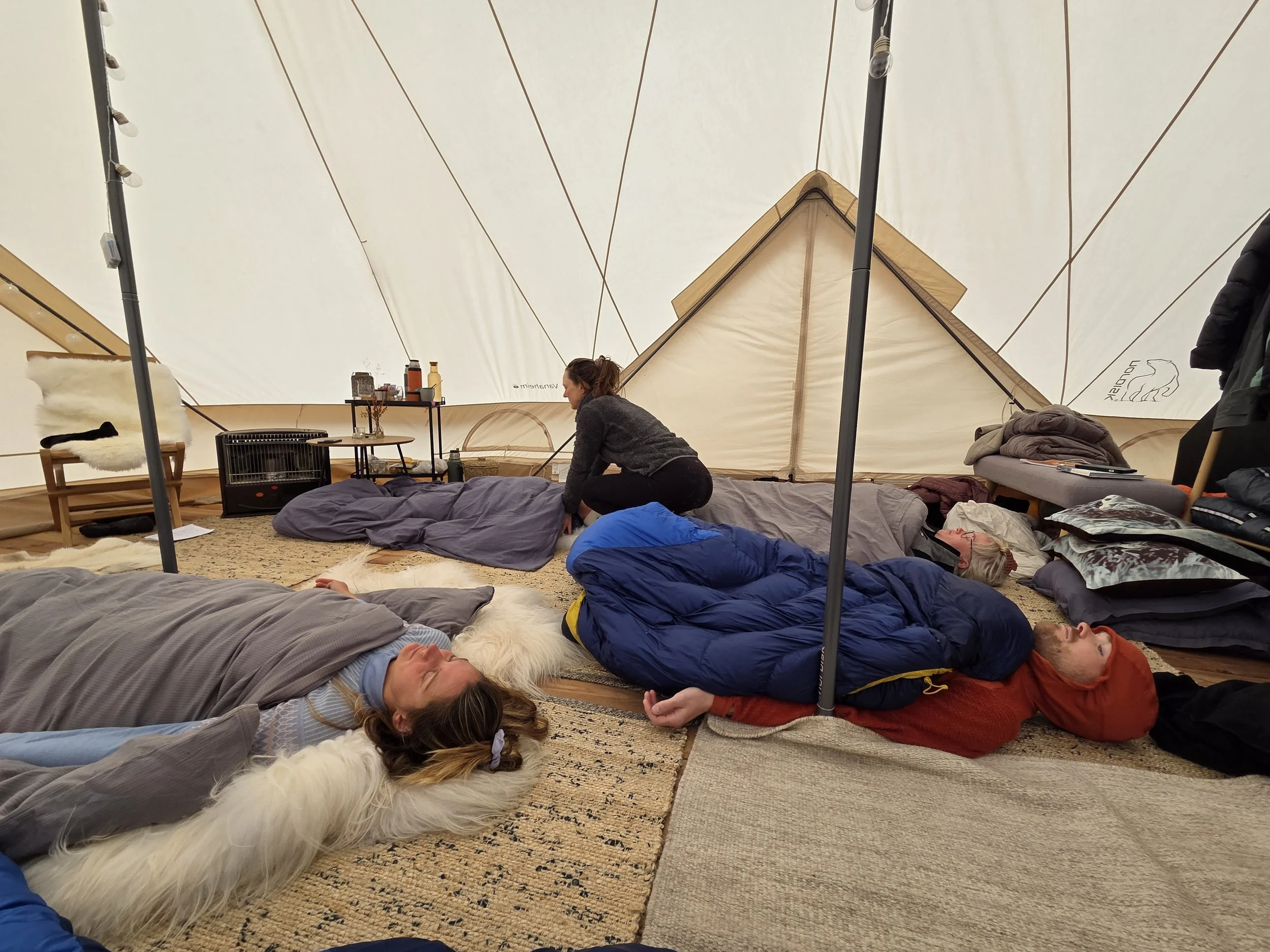 Inside a large tent with people meditating on the floor and one person giving hands on assist. There are various pillows, blankets, and items on a bench and nearby surfaces. Glamping tents in Greenland