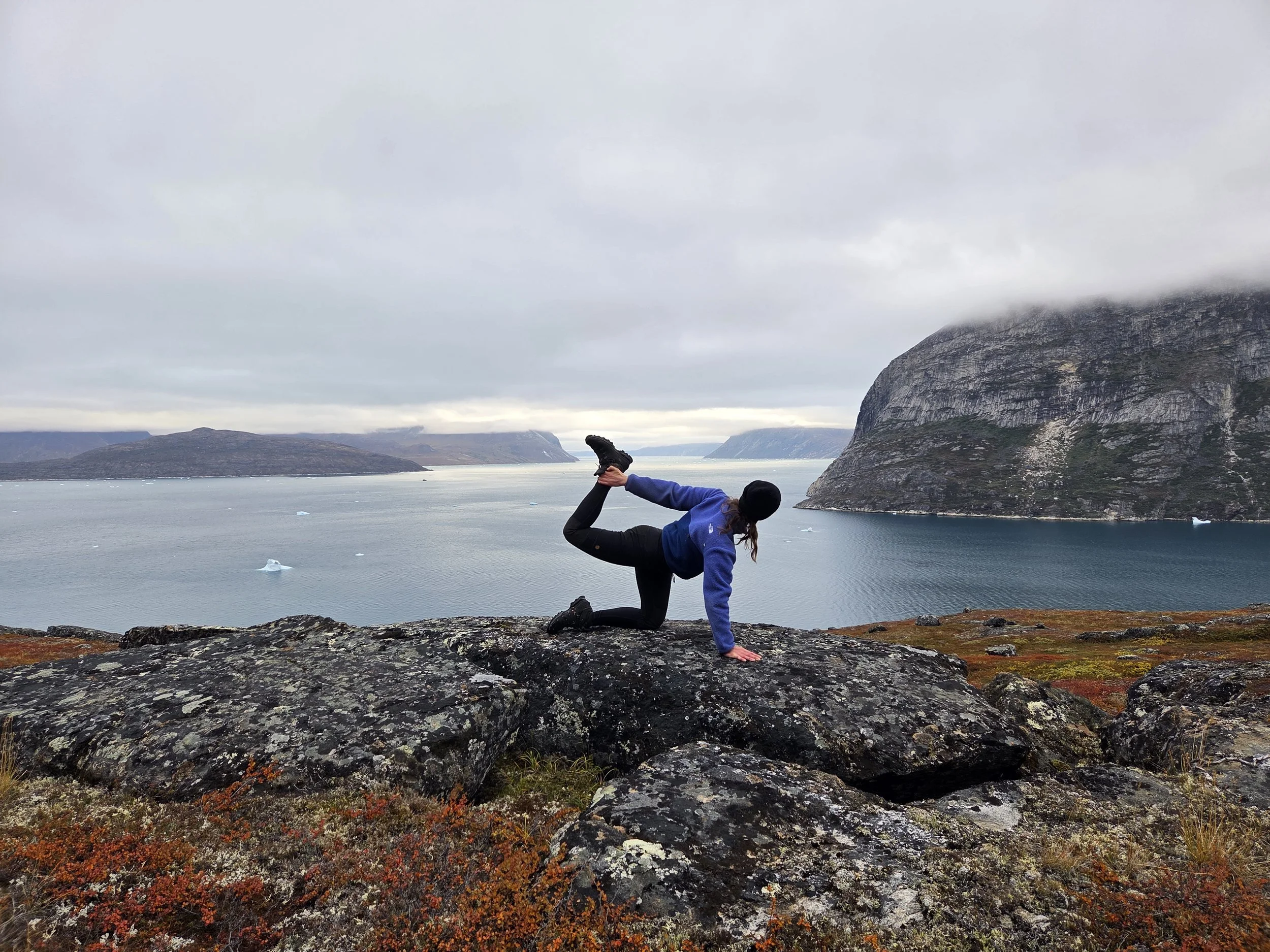 Person in athletic clothing performing a yoga pose on rocky terrain overlooking a fjord with icebergs, mountains, and overcast sky in the background in Greenland