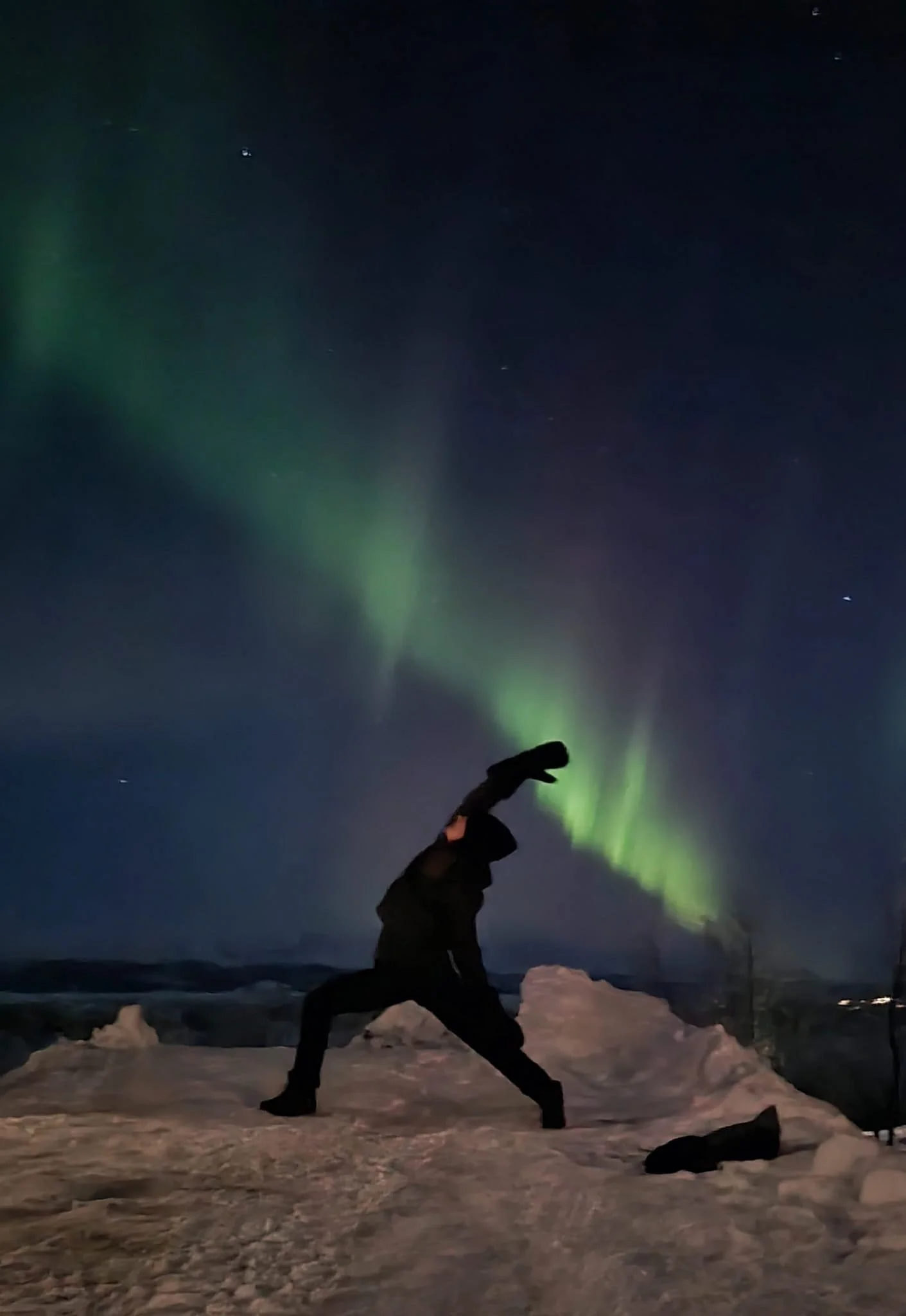 Person dressed in black, performing a yoga pose, reversed warrior, on snow under the Northern Lights at night in Sweden.