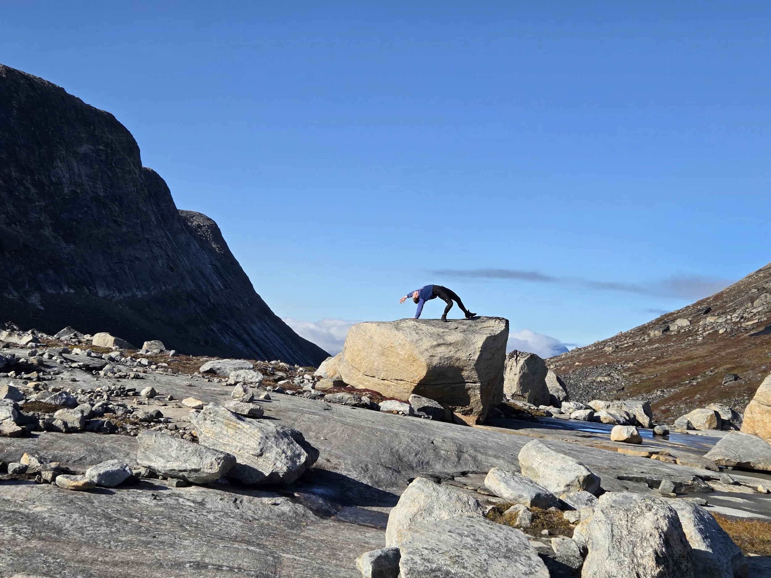 Person in black pants and blue top performing yoga pose on a large rock in a mountainous landscape with large boulders and clear blue sky in Greenland