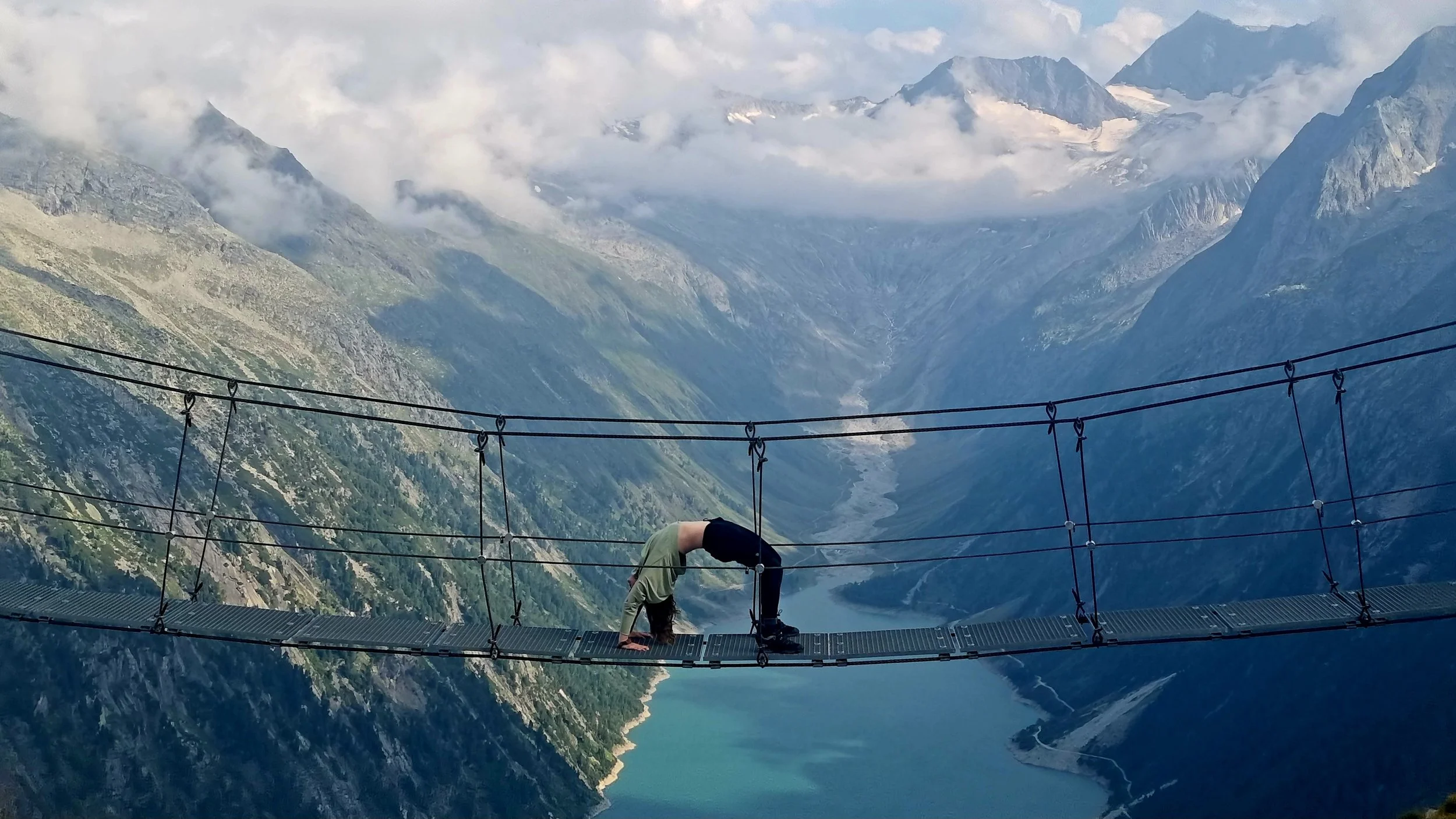 Person in black shirt and pants performing a backbend on a suspension bridge over a deep mountain valley with a river, surrounded by high mountains with some snow and cloud cover.
