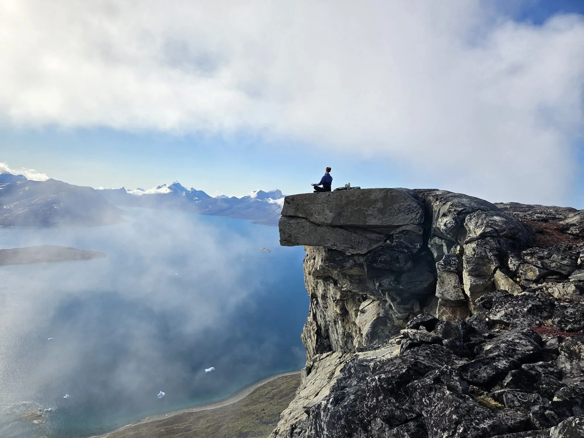 A person practicing meditation or yoga on a rock ledge overlooking a mountain lake with mist and snow-capped peaks in the background in Greenland outdoorretreat