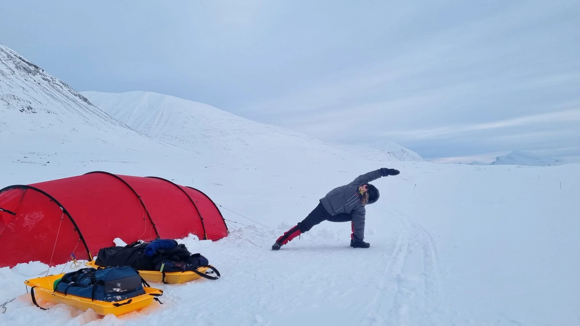 A person in winter gear doing a extended side angel yogapose, in a snowy landscape next to a red tent and sleds, with snow-covered mountains in the background In Sampi, Lapland in Sweden Kunsleden. Winterhiking and camping