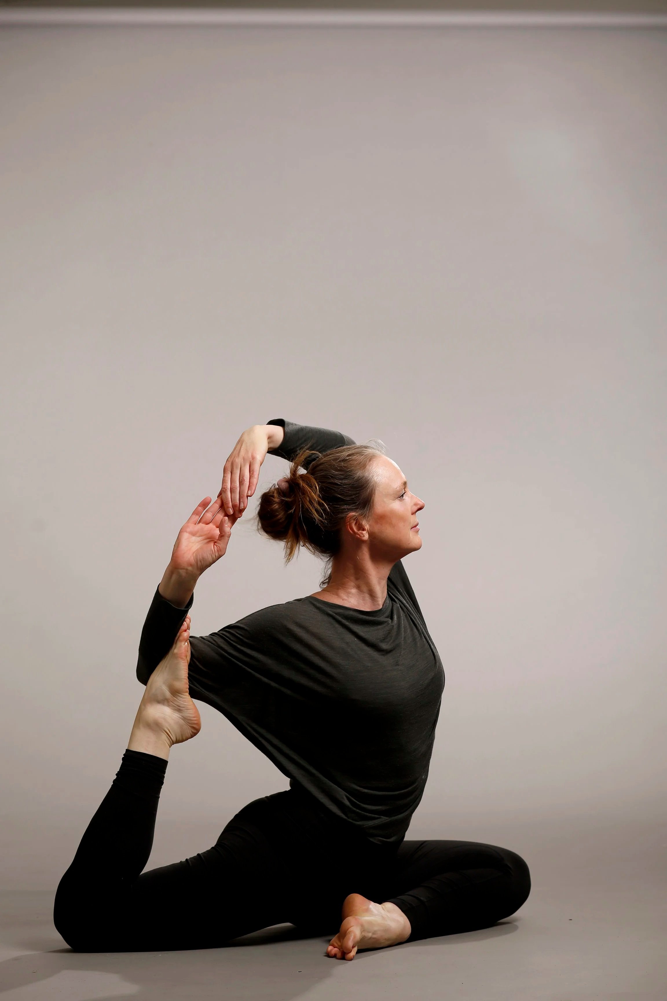 A woman practicing yoga in a seated pose with her left leg bent and foot on the ground, her right leg extended upward, and her right hand holding her right foot while her left arm is extended overhead.
