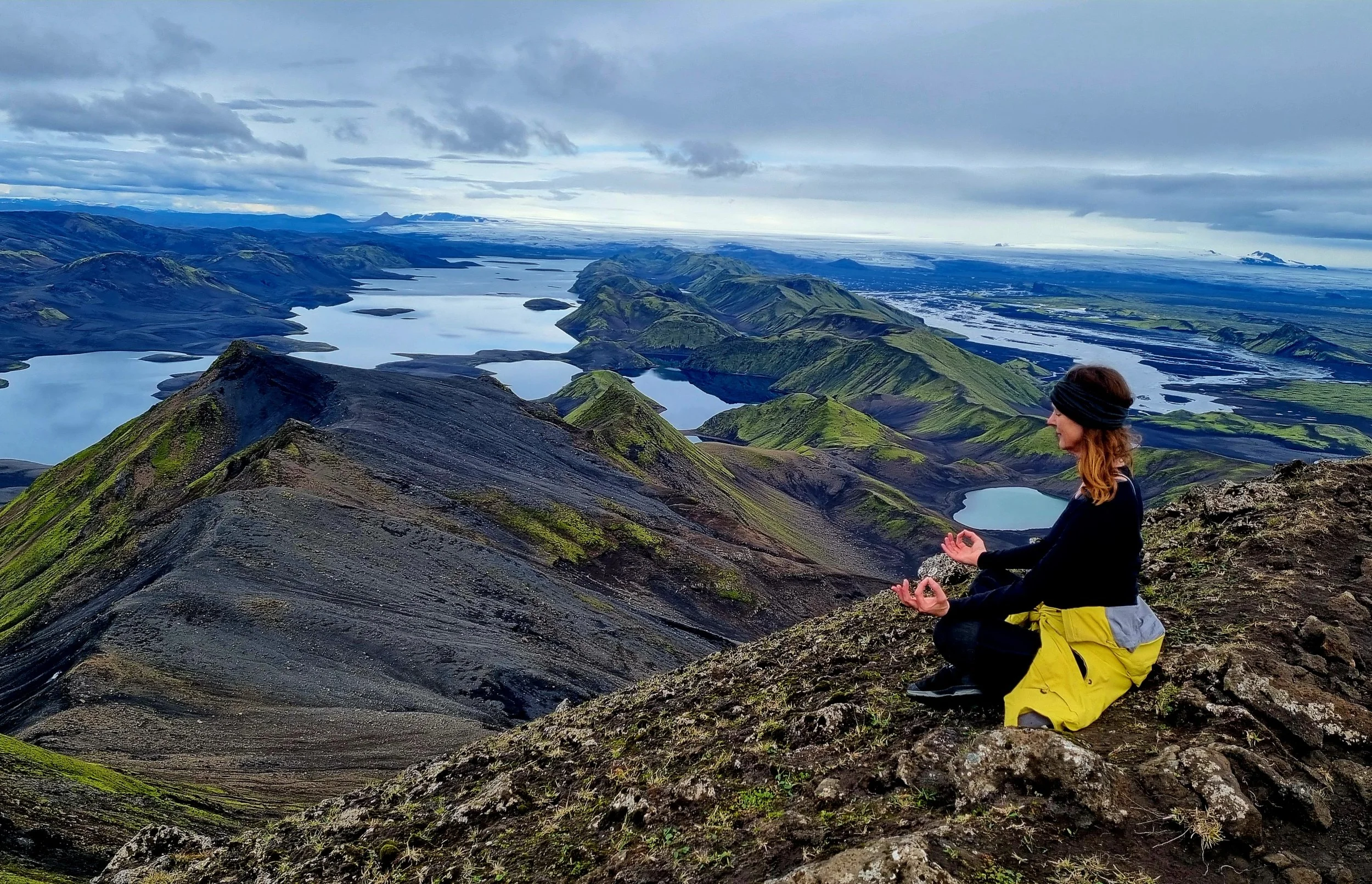 A woman meditating on a mountain summit overlooks a landscape of green hills, lakes, and mountains under a cloudy sky.