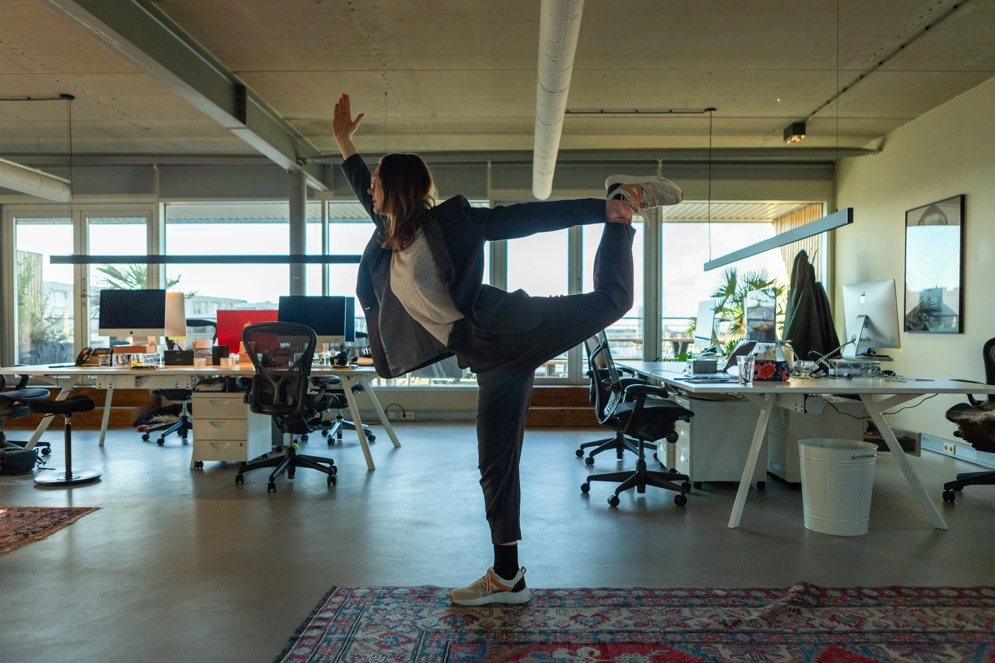 Woman practicing yoga in an office with desks, chairs, computers, and large windows in Amsterdam.