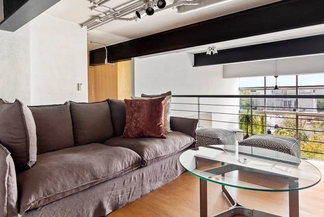 Modern living room with a gray sofa, a glass coffee table, and large windows with blinds, overlooking city buildings.