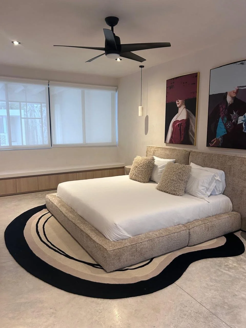Modern bedroom with a low beige bed, white linens, two fluffy beige pillows, and two white pillows, on a black and beige oval rug, with artwork on the wall, large windows with white blinds, a black ceiling fan, and a pendant light.