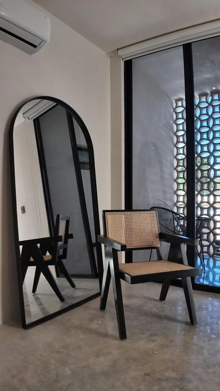 A modern room with a large arched mirror, a chair with a wooden frame and woven back and seat, a glass sliding door with a decorative patterned screen or wall behind it, and an air conditioning unit on the wall.