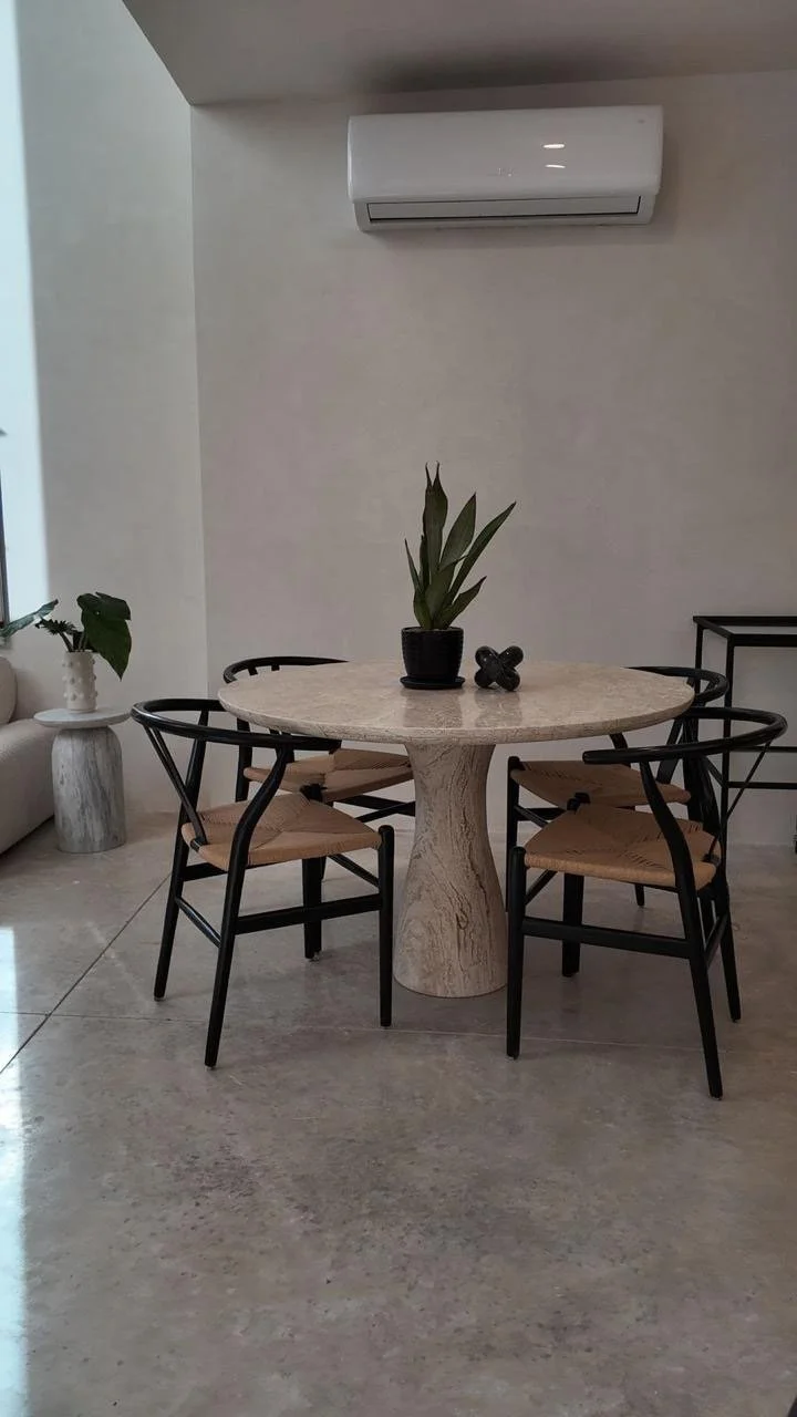 A round beige marble table with four black and wood chairs in a modern dining area, decorated with a potted plant and a small black decorative object, with air conditioning and minimalist decor.