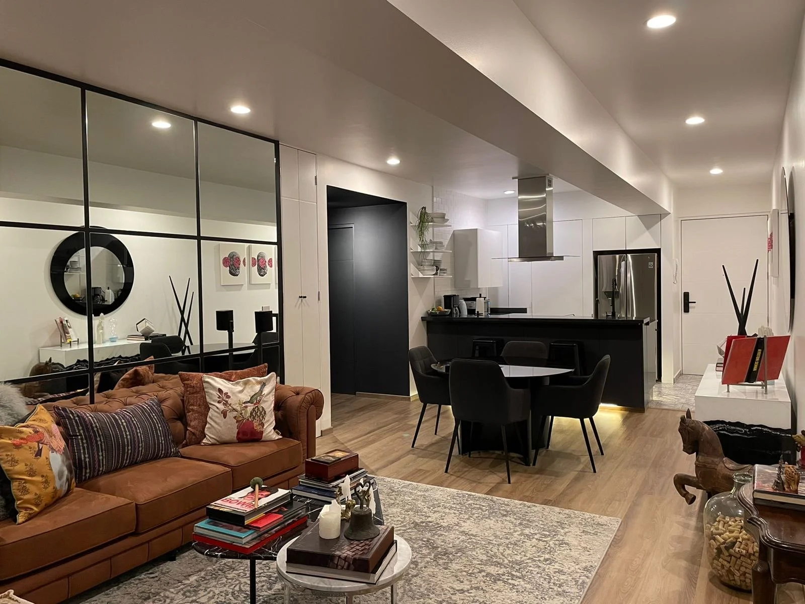 Living room with a brown leather sofa, pillows, a coffee table with books and decorative items, a dining area with a round table and black chairs, kitchen with white cabinets, stainless steel refrigerator, black countertop, and modern lighting with r