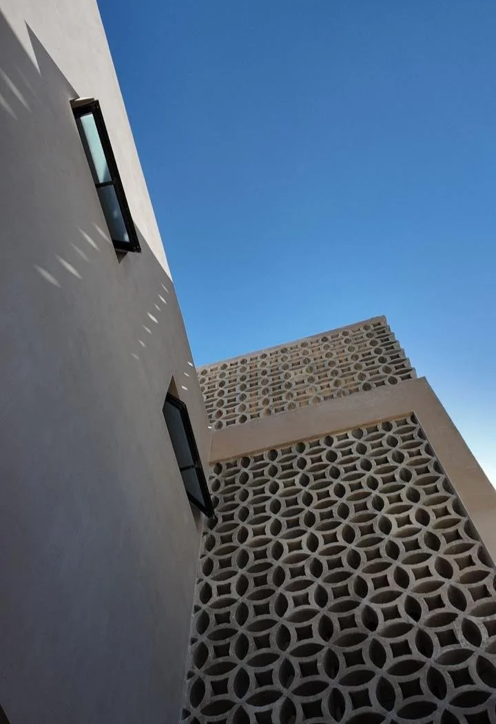 A low-angle view of a white building with two small windows, shadows cast by the window frames, and a decorative wall with geometric circular patterns, under a clear blue sky.