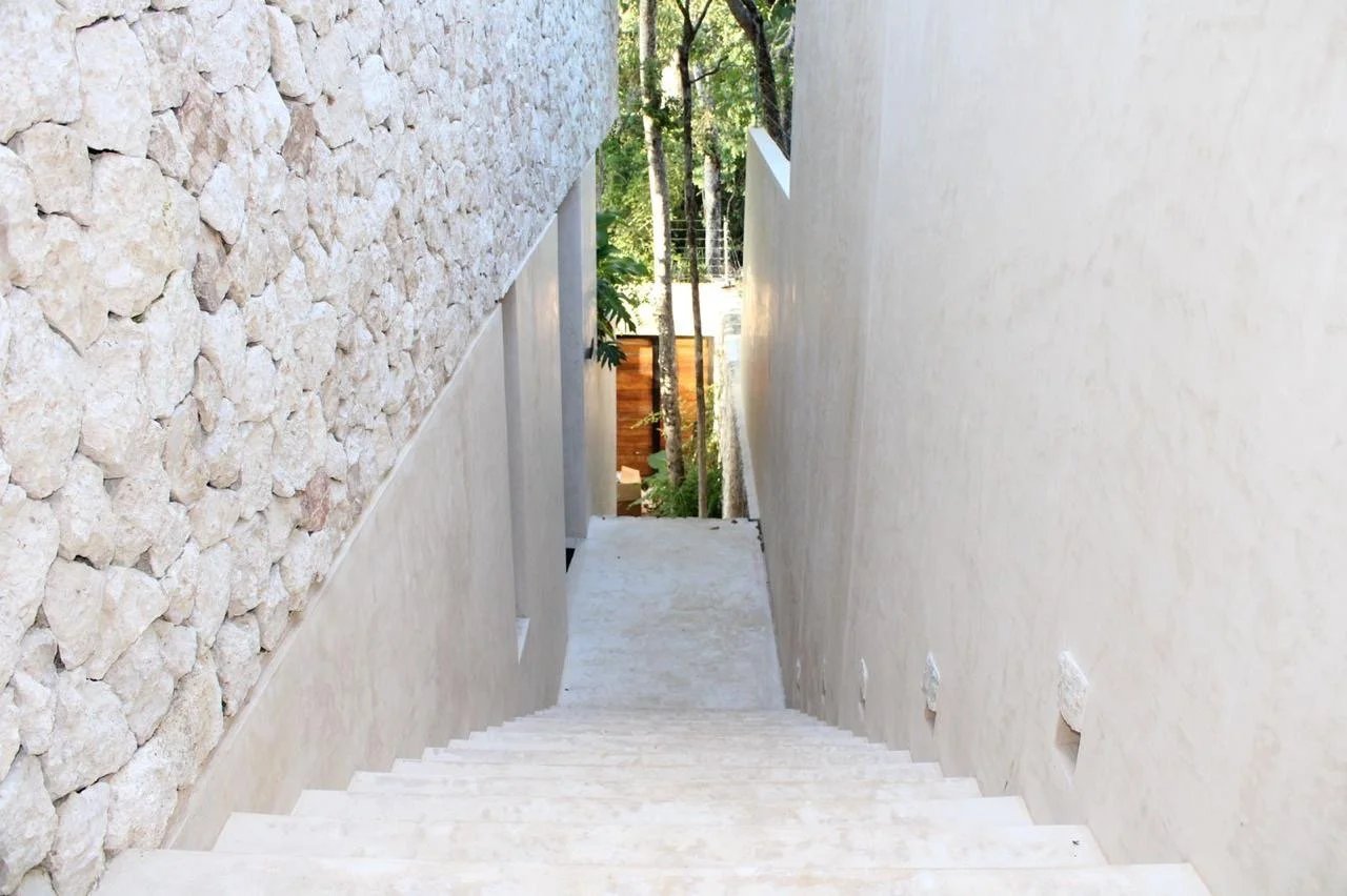 A view of a white outdoor staircase between two walls, one made of stone and the other smooth, leading downwards to a green, wooded area.