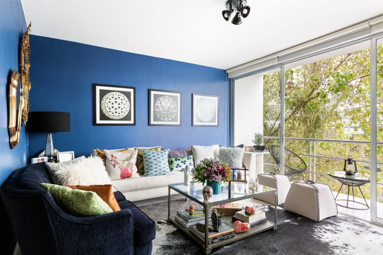Modern living room with a blue accent wall, colorful throw pillows on a white sofa, a black sofa with green and orange pillows, and large glass sliding doors leading to a balcony with outdoor seating and green tree view.