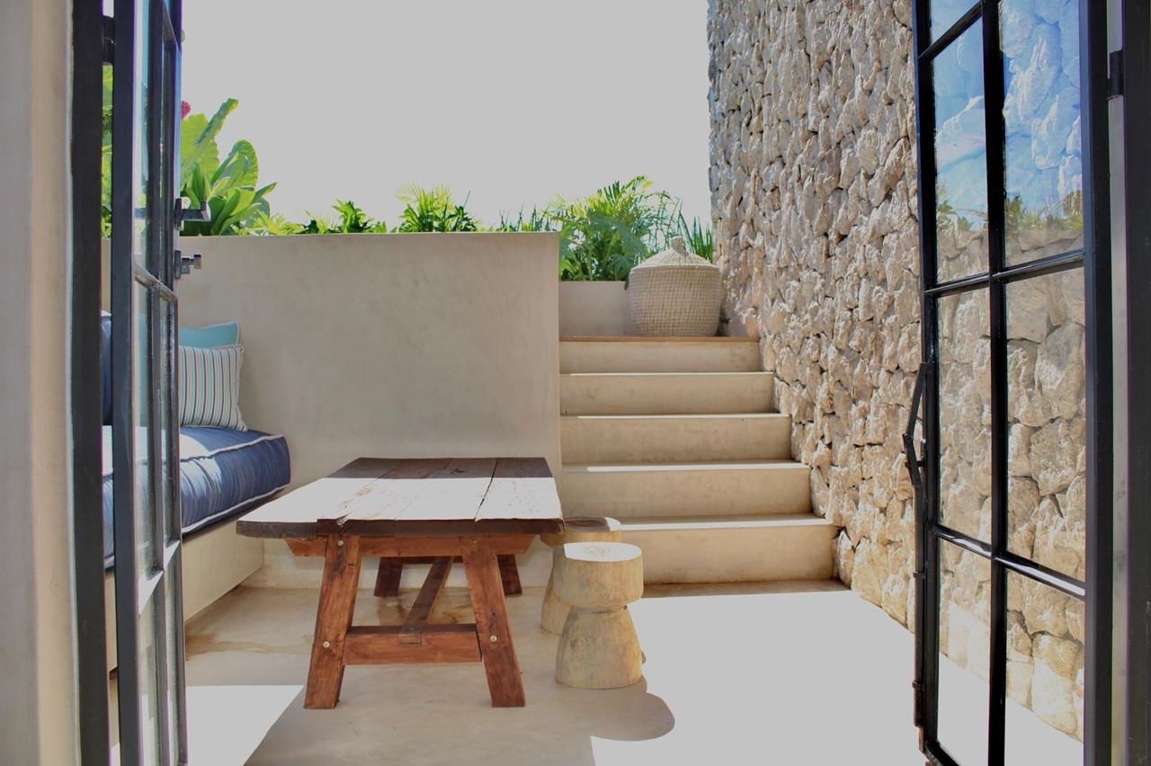 An outdoor patio area featuring a wooden bench, a small wooden stool, stone steps, a beige wall, a stone wall, and a black metal door with glass panels, with plants visible in the background.