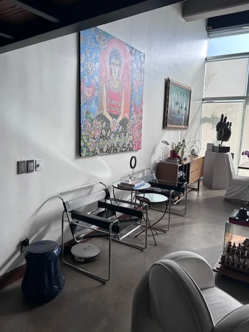 Modern interior space with abstract wall art, a seating area with black and chrome chairs, a round side table, and decorative items including a helmet sculpture and a bookshelf, with large windows letting in natural light.