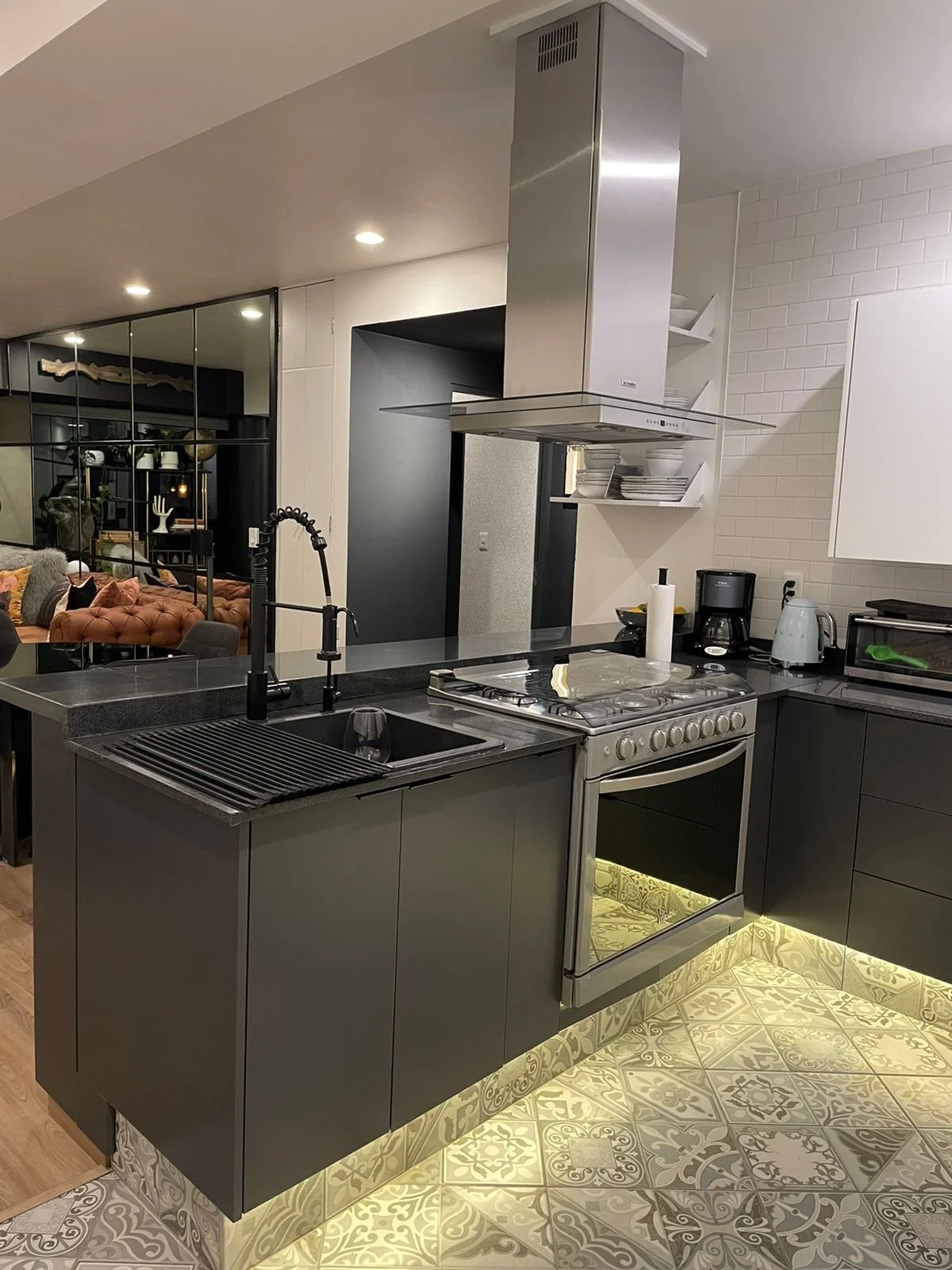 Modern kitchen with black cabinets, stainless steel appliances, a black sink, and patterned tile flooring, open to a living room with colorful seating and decorative items.