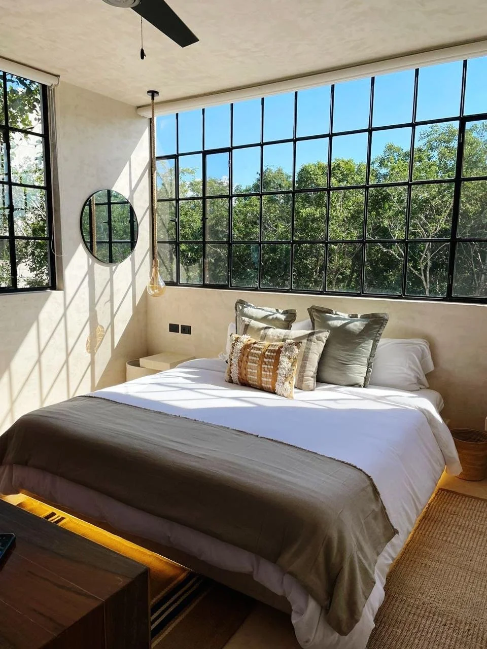 A bedroom with large black-framed windows allowing natural sunlight and a view of green trees outside. The bed has neutral-colored pillows and a white bedspread, with a beige throw blanket at the foot. A small nightstand is beside the bed, and a mirr
