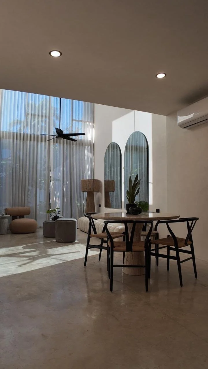 Modern living room with large windows, sheer curtains, a ceiling fan, a round dining table with four chairs, decorative mirrors, potted plants, and stylish lighting.