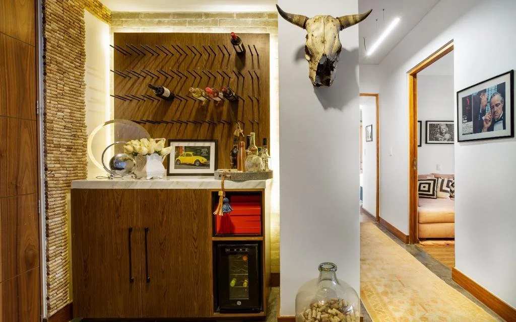 Interior of a home with a bar area, white walls, a mounted cow skull, decorative bottles, framed pictures, and a hallway with a beige rug leading to rooms.