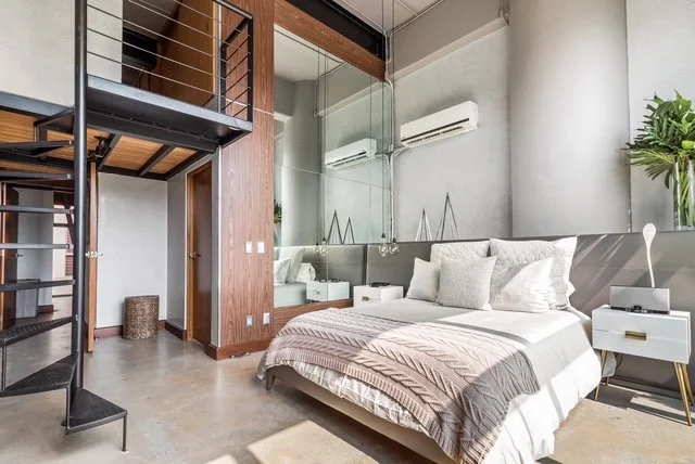 Modern bedroom with a large bed, white pillows, and a beige blanket. A nightstand with a lamp and decorative object, a large potted plant, and an air conditioning unit on the wall. Glass partition with a mirror, wooden accents, and metal spiral stair