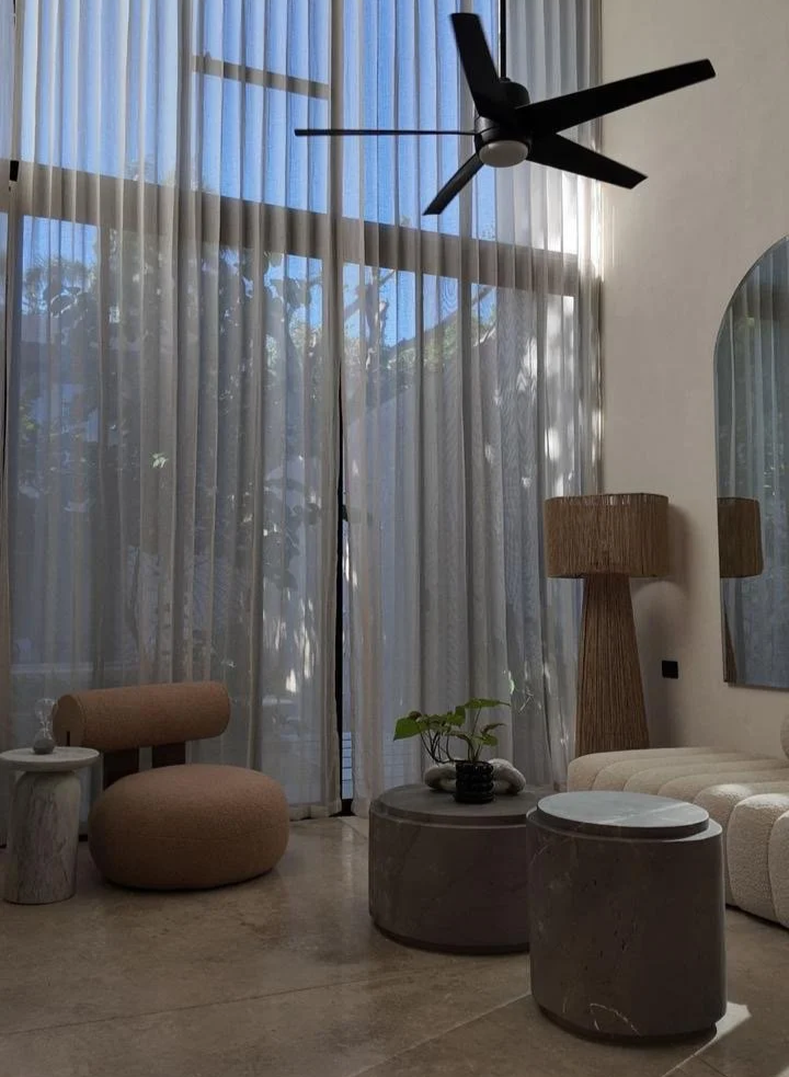 Living room with large sheer curtains, a ceiling fan, a beige rounded chair, a small marble side table, a decorative plant, a large wooden floor lamp, and modern marble coffee tables.