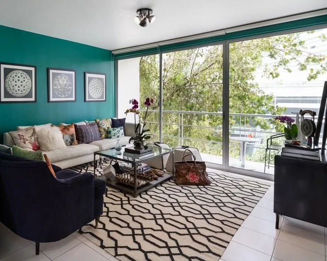 Living room with teal accent wall, white sofa with multiple pillows, navy armchair, glass coffee table, black and white geometric patterned rug, large glass sliding doors with greenery outside, framed geometric artwork, and various decorative items.