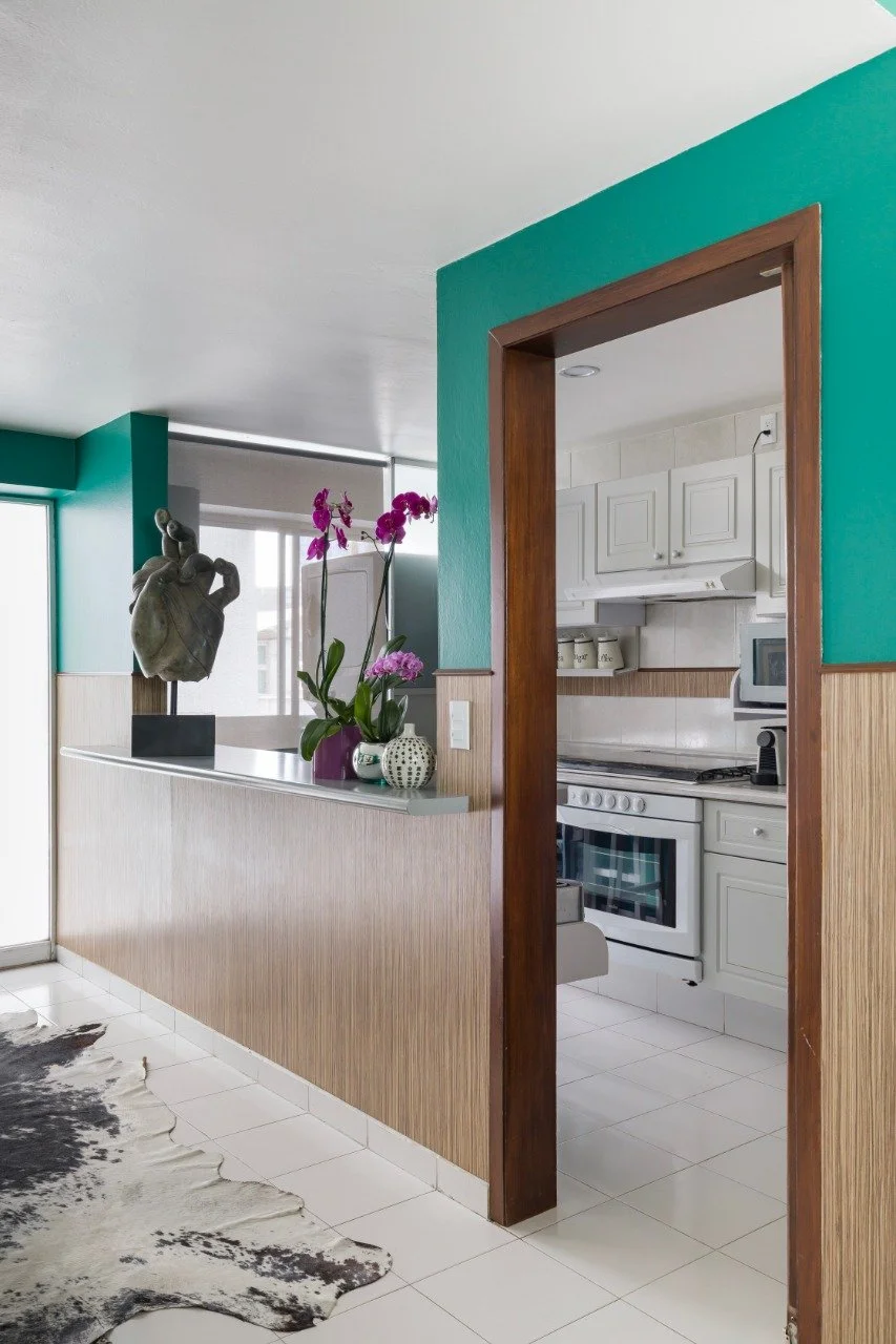 Interior view of a kitchen with white cabinets, white appliances, and a teal wall, seen through a doorway. There is a purple orchid plant and decorative vases on a half-wall that separates the kitchen from the adjacent room. The kitchen floor is tile