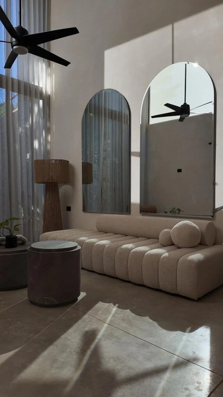 Modern living room with a beige textured sofa, circular pillows, and two arched mirrors on a light-colored wall. A wooden floor lamp with a cylindrical shade stands next to sheer curtains, and a black ceiling fan hangs from the ceiling. Sunlight crea