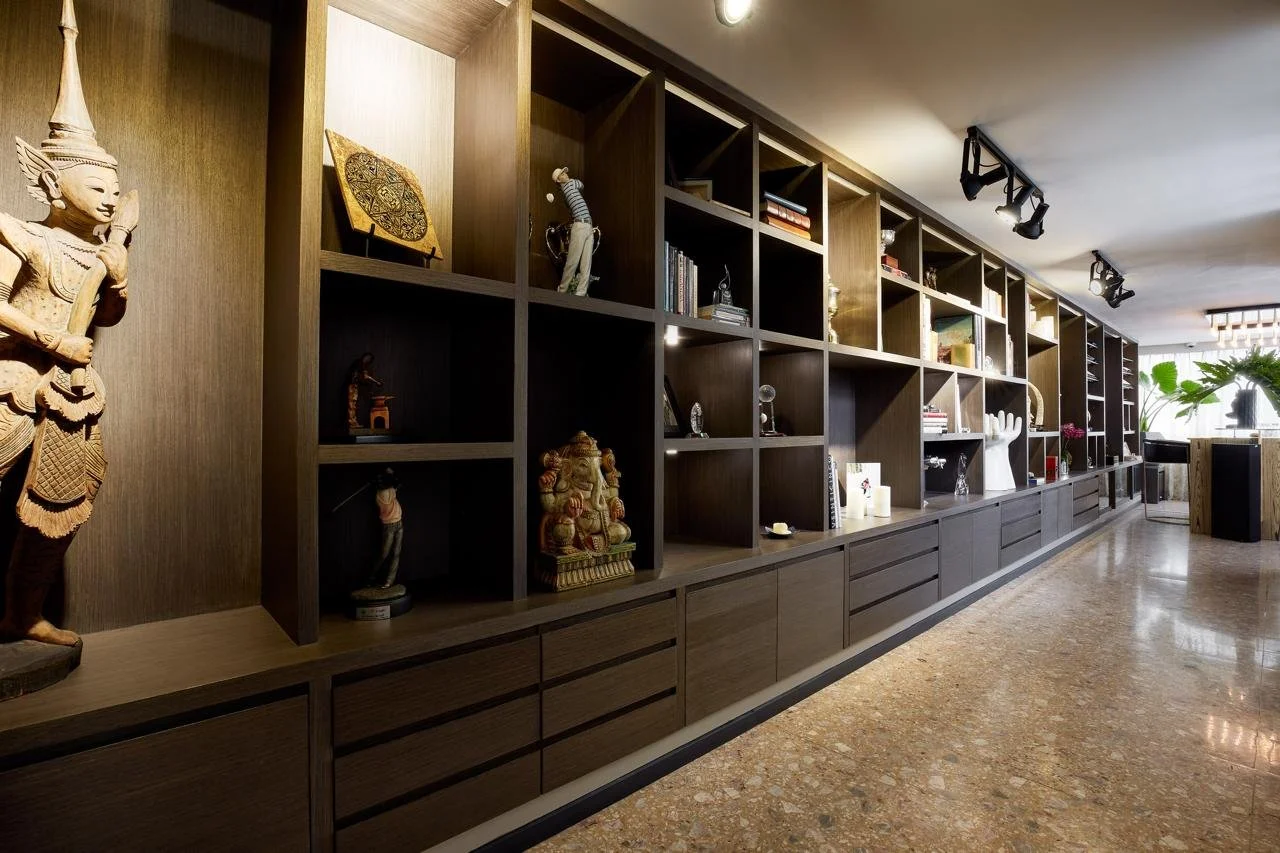 A long wooden display shelf with open cubbies holding various decorative items, sculptures, and books in a well-lit room with a terrazzo floor and some plants near the window.