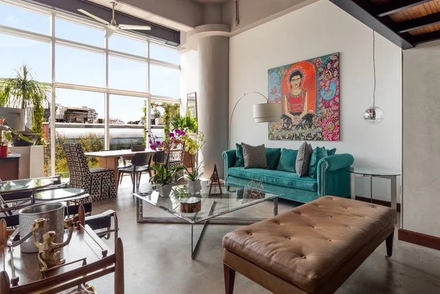 Interior of a modern living room with large windows, a teal sofa, colorful artwork, a glass coffee table, and various plants.