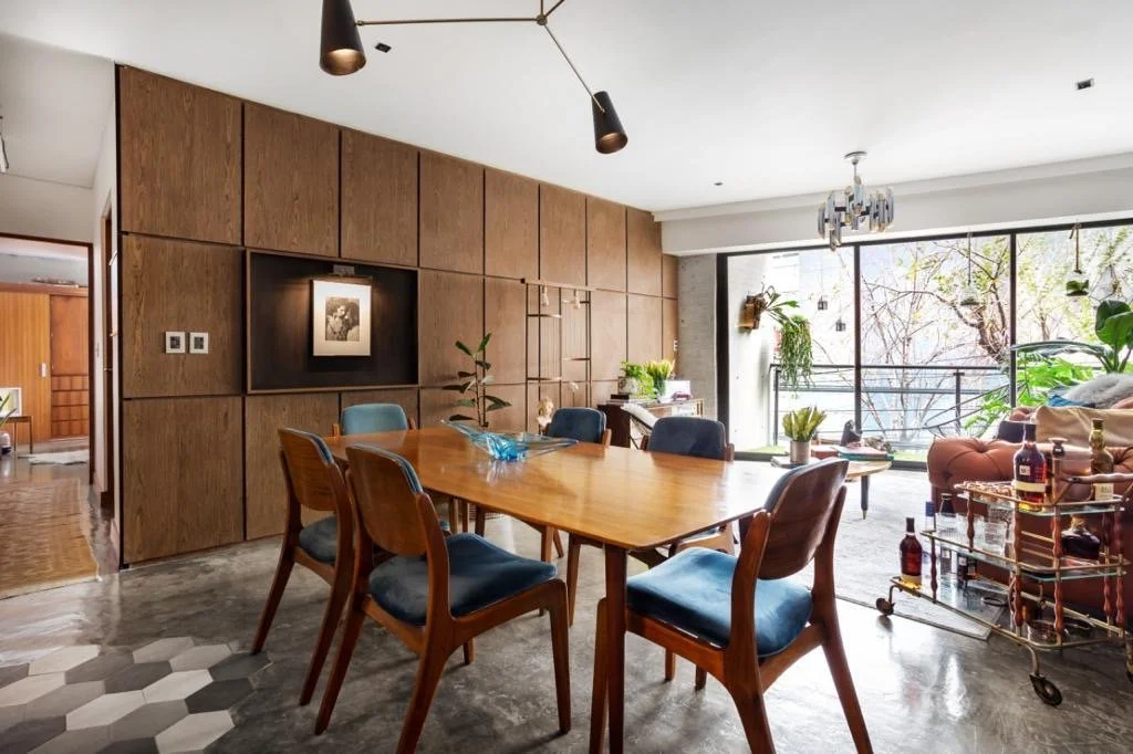 A living room and dining area with a wooden dining table and chairs, a large window with a balcony, potted plants, a sofa, and a bar cart in a modern, well-lit apartment.