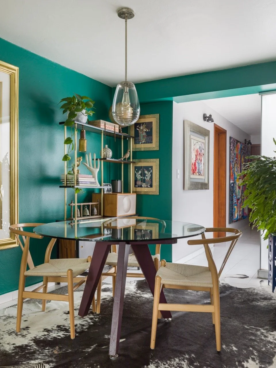 Dining area with a black glass-top table, two light wooden chairs with woven seats, a green accent wall, modern shelves, houseplants, art frames, and a cowhide rug.
