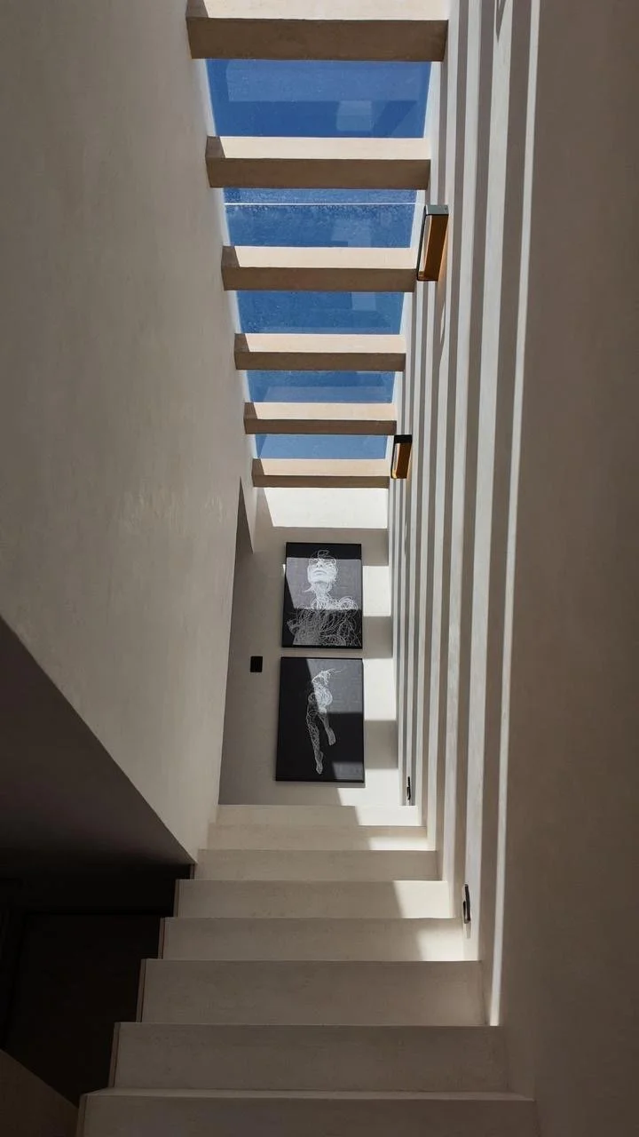Interior shot of a modern staircase with open wooden steps, natural sunlight streaming in, and artwork hanging on the wall at the bottom of the stairs, with a view of a blue sky outside.