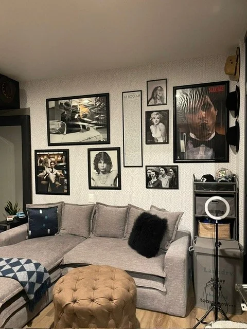 Living room with a gray sectional sofa, a black pillow, and a brown tufted ottoman. The wall has several framed black-and-white and color photos, including musicians and actors. There's a TV on the left, a small shelf with electronic devices and a ri